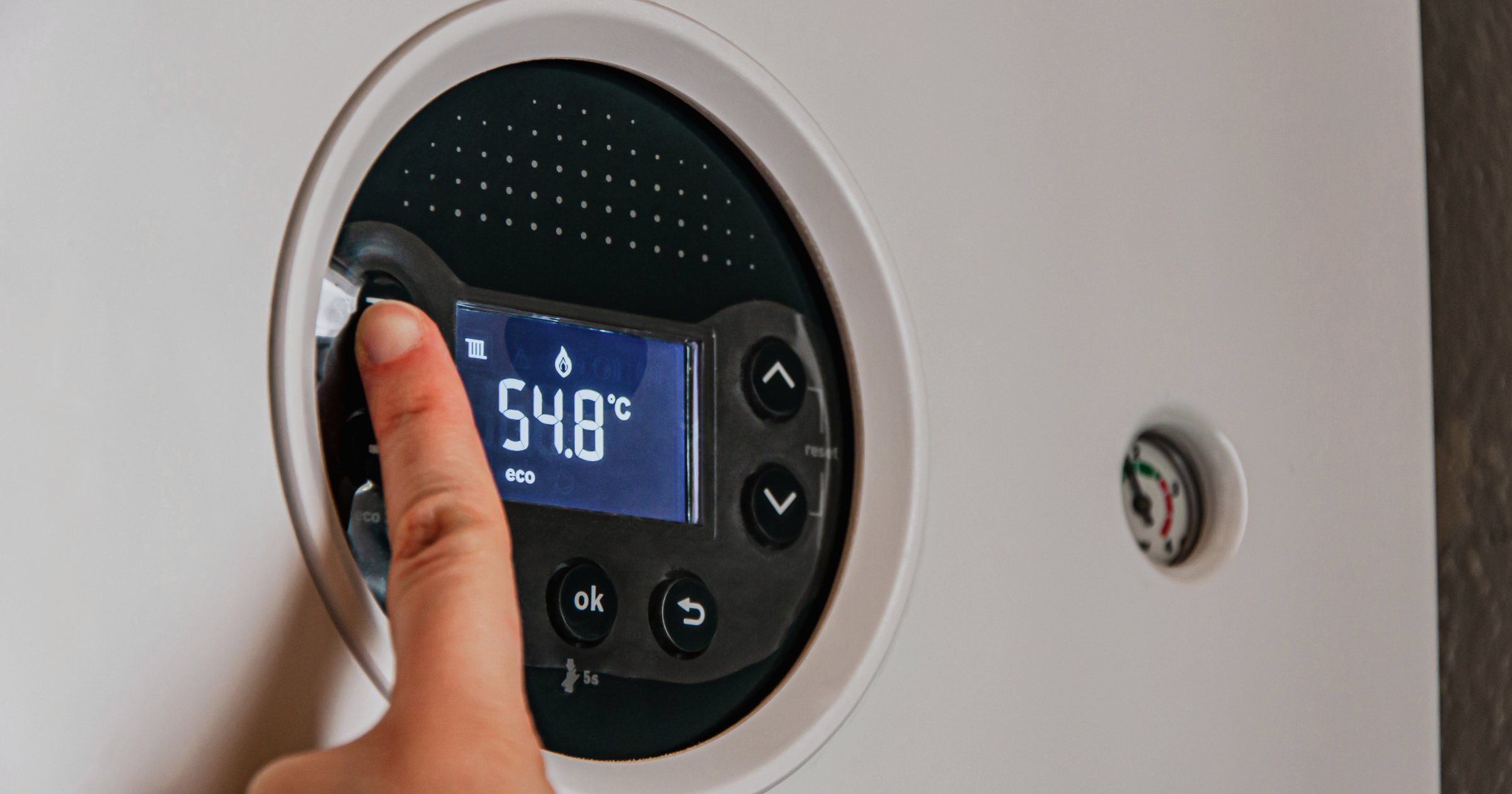 Mâna unei persoane reglează panoul de comandă digital al unei centrale termice pe gaz montată pe perete; pe afișaj apare temperatura 54,8°C și butoanele de navigare